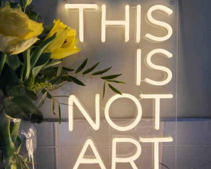 Photo of LED Neon Sign saying THIS IS NOT ART next to a vase of yellow flowers by Love Inc.